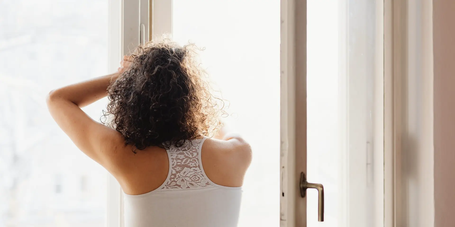 Femme de dos aux cheveux bouclés, devant une fenêtre lumineuse